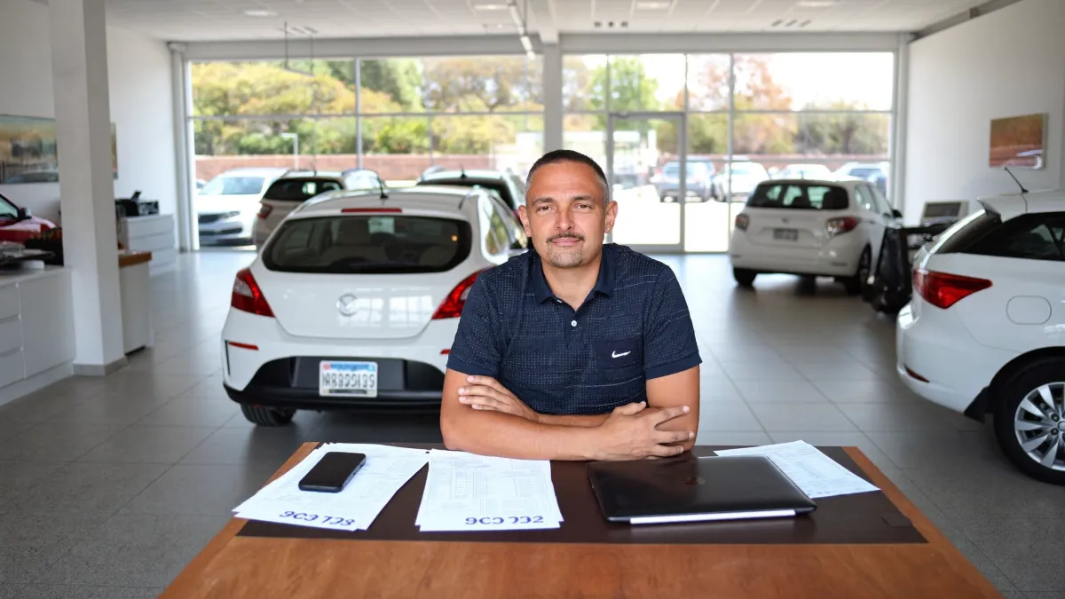 Dono de revenda de carros conferindo documentos de veículos no notebook, com o pátio da loja ao fundo